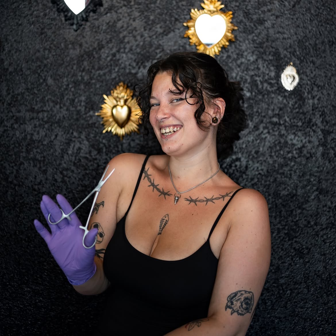 Jade, tout sourire, pince de piercing à la main devant le mur d'ex-voto dorés du salon